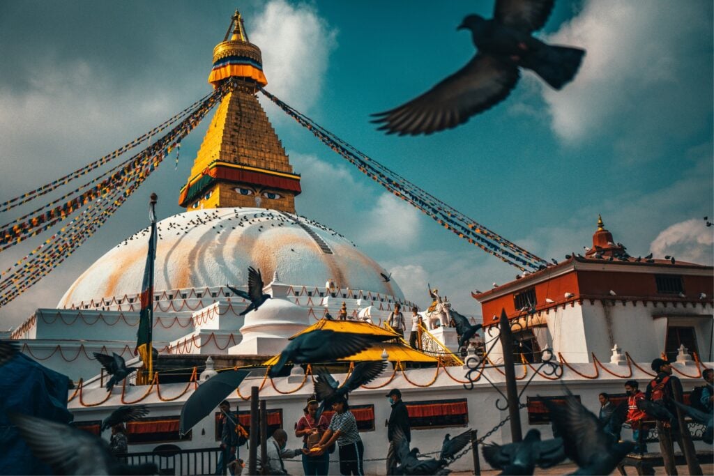 Pigeons taking flight in front of Boudhanath Stupa's golden spire and painted eyes, with visitors feeding birds below