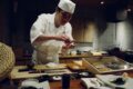 Japanese sushi chef shaping nigiri by hand at a counter, with a sashimi display case and traditional knives