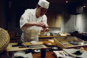Japanese sushi chef shaping nigiri by hand at a counter, with a sashimi display case and traditional knives