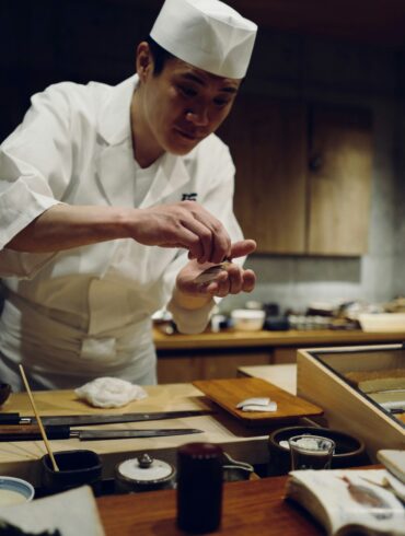 Japanese sushi chef shaping nigiri by hand at a counter, with a sashimi display case and traditional knives