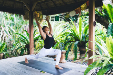 Woman practising a low lunge pose on a yoga mat in an open-air garden shala decorated with Balinese prayer flags