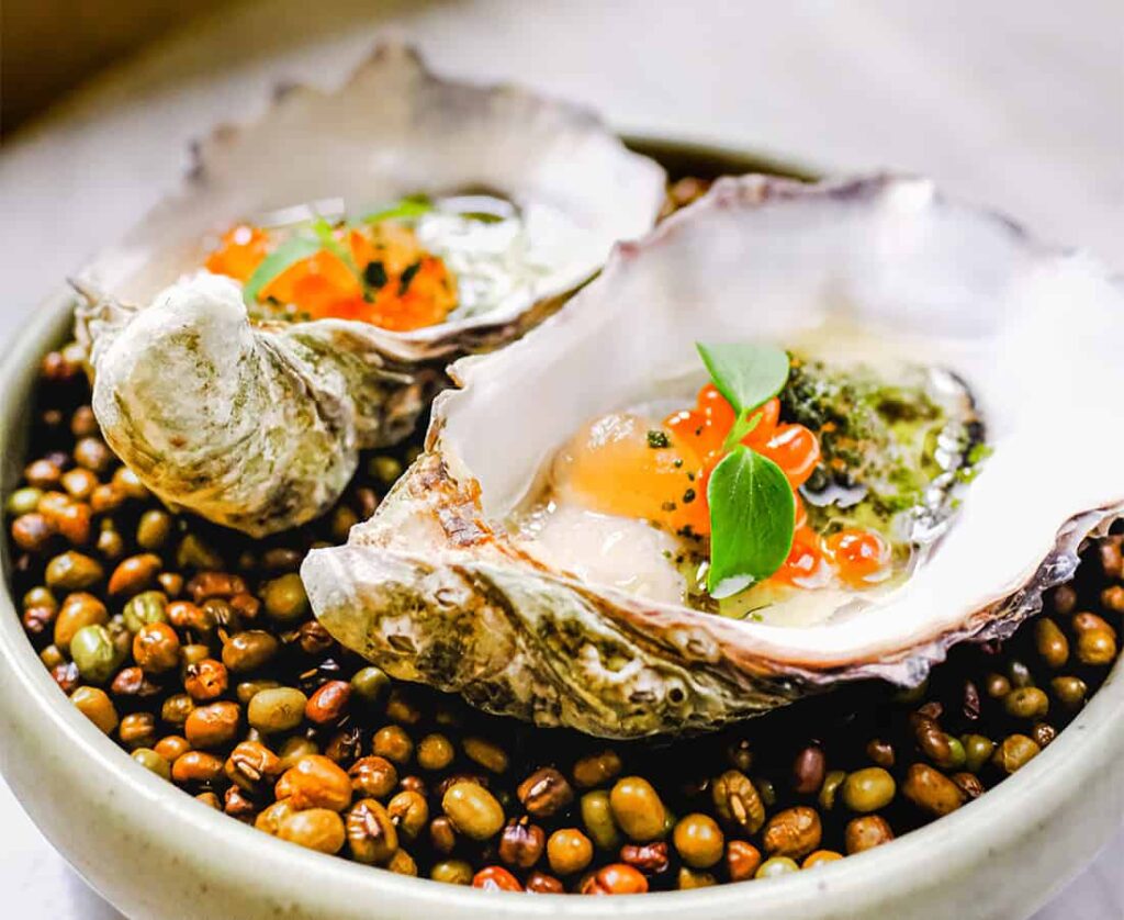 19:58Claude responded: Two oysters served on the half shell, nestled in a bed of mung beans.Two oysters served on the half shell, nestled in a bed of mung beans. Each is topped with salmon roe, microgreens, and a green herb sauce, with one finished with black caviar.