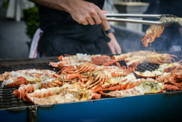 Chef grilling fresh lobsters and crabs on a large outdoor barbecue grill, with smoke rising.
