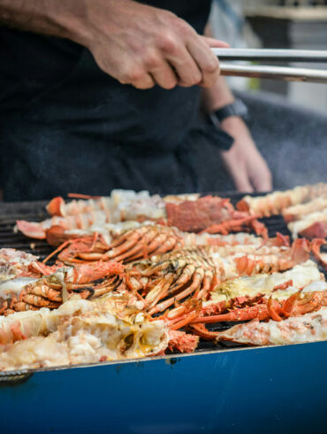 Chef grilling fresh lobsters and crabs on a large outdoor barbecue grill, with smoke rising.