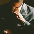 Close-up of a man in a tailored charcoal suit adjusting his tie, hand resting on the jacket's lapel