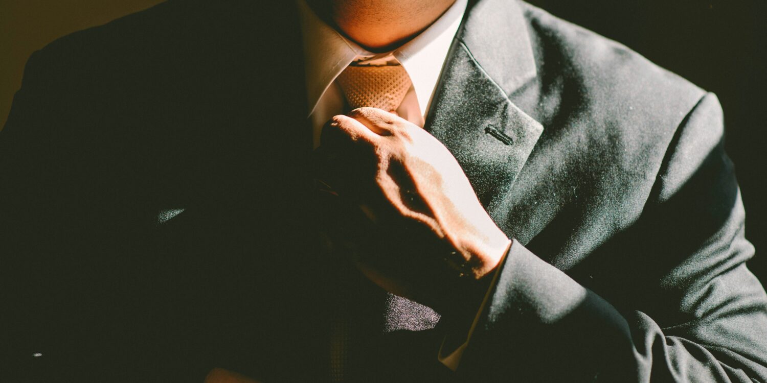 Close-up of a man in a tailored charcoal suit adjusting his tie, hand resting on the jacket's lapel