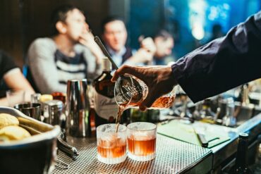 Bartender pouring craft cocktails into rocks glasses at a busy cocktail bar, showcasing mixology techniques and vibrant nightlife atmosphere in Singapore.