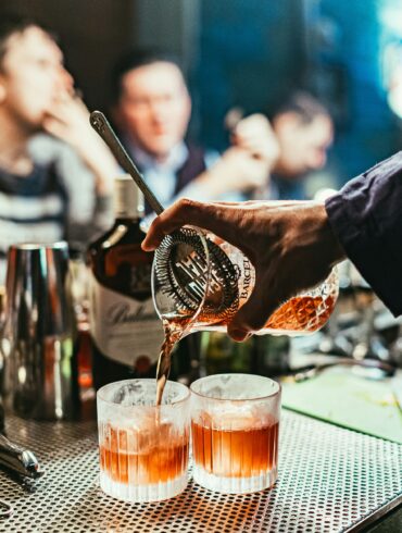 Bartender pouring craft cocktails into rocks glasses at a busy cocktail bar, showcasing mixology techniques and vibrant nightlife atmosphere in Singapore.