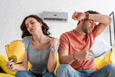 Pretty woman with hand fan and handsome man with newspaper suffering from heat at home