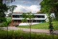 Exterior view of AIR CCCC at Dempsey Hill, a modern white two-storey restaurant with floor-to-ceiling glass windows, set on a manicured lawn and flanked by lush tropical greenery, with a wooden boardwalk leading up to the deck.