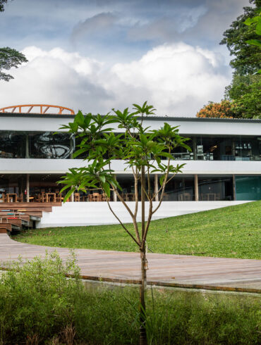 Exterior view of AIR CCCC at Dempsey Hill, a modern white two-storey restaurant with floor-to-ceiling glass windows, set on a manicured lawn and flanked by lush tropical greenery, with a wooden boardwalk leading up to the deck.