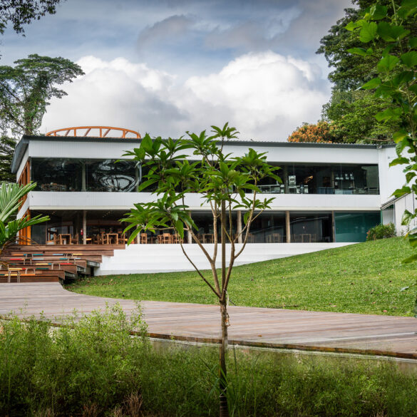 Exterior view of AIR CCCC at Dempsey Hill, a modern white two-storey restaurant with floor-to-ceiling glass windows, set on a manicured lawn and flanked by lush tropical greenery, with a wooden boardwalk leading up to the deck.