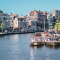 Canal boats docked along an Amsterdam canal lined with historic gabled buildings and a passing tram