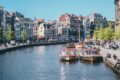 Canal boats docked along an Amsterdam canal lined with historic gabled buildings and a passing tram