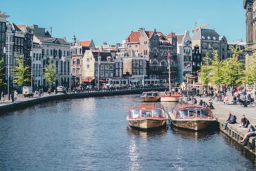 Canal boats docked along an Amsterdam canal lined with historic gabled buildings and a passing tram