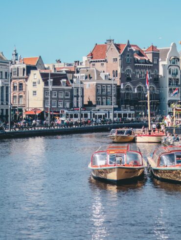 Canal boats docked along an Amsterdam canal lined with historic gabled buildings and a passing tram