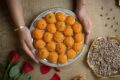 Hands holding a silver tray of orange laddu topped with pistachio and almond slivers, a Deepavali sweet, beside a bowl of pistachios.