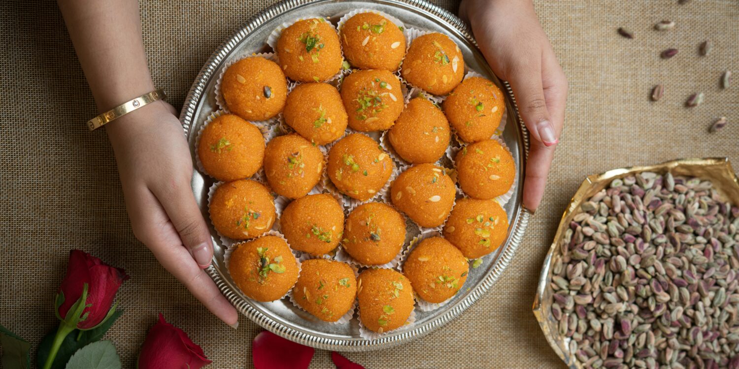 Hands holding a silver tray of orange laddu topped with pistachio and almond slivers, a Deepavali sweet, beside a bowl of pistachios.