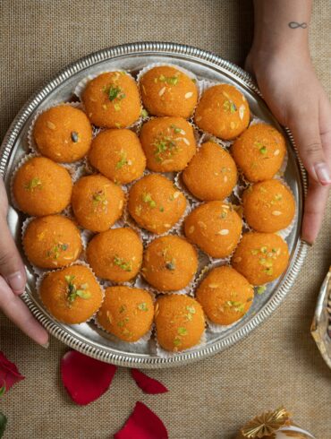 Hands holding a silver tray of orange laddu topped with pistachio and almond slivers, a Deepavali sweet, beside a bowl of pistachios.