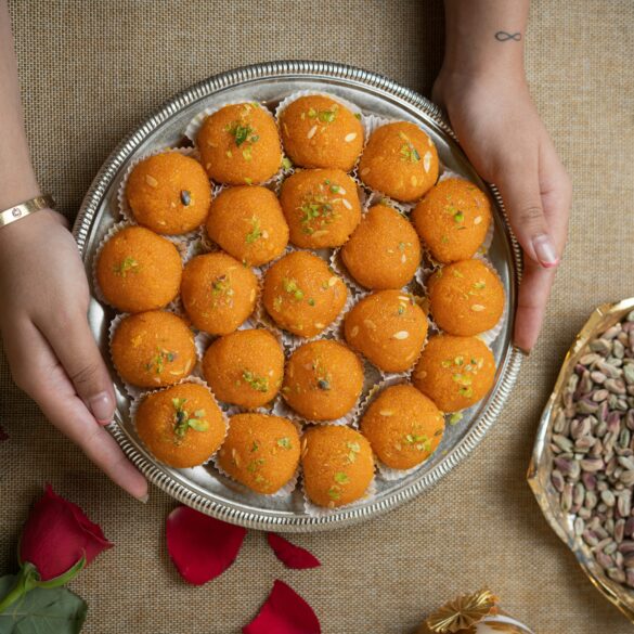 Hands holding a silver tray of orange laddu topped with pistachio and almond slivers, a Deepavali sweet, beside a bowl of pistachios.