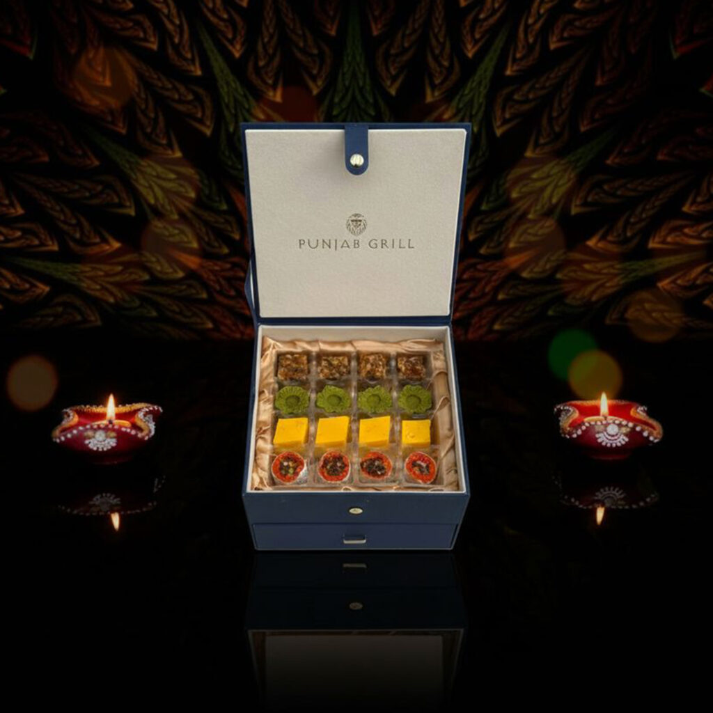 Punjab Grill Khushi Box in navy, opened to show 16 mithai in four rows, flanked by lit diyas.