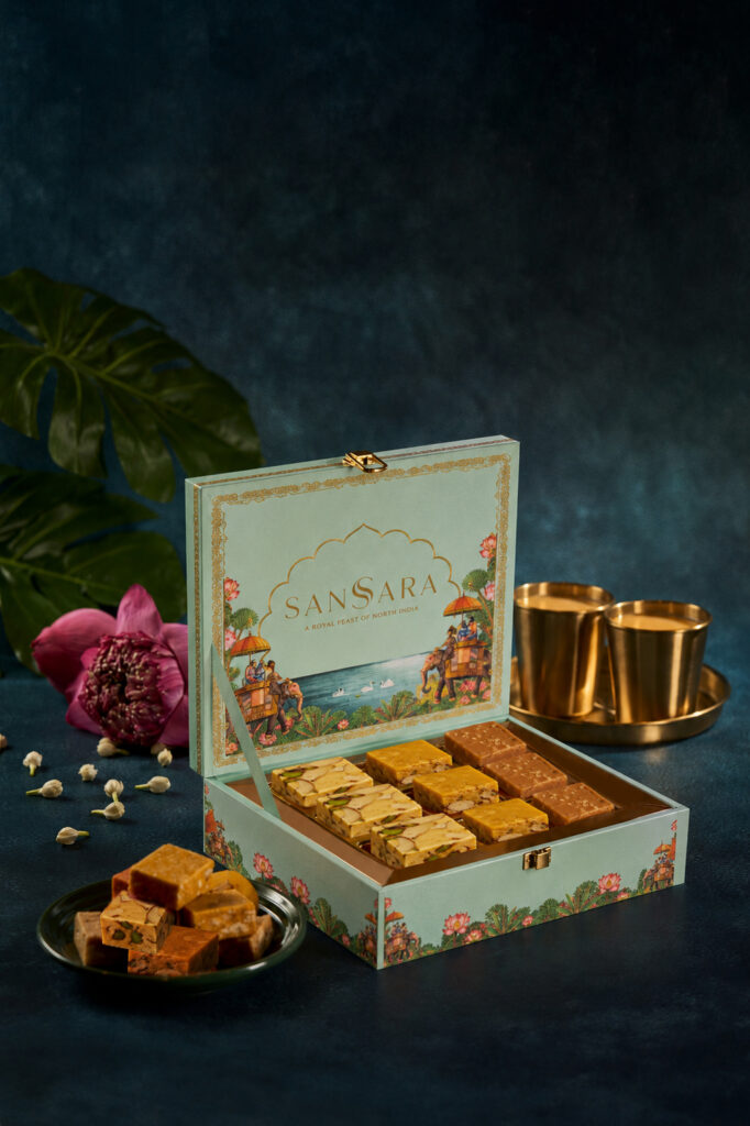 SanSara Diwali Mithai Gift Box with nine pieces of soan papdi, Mysore pak and malt burfi, beside brass cups of chai.