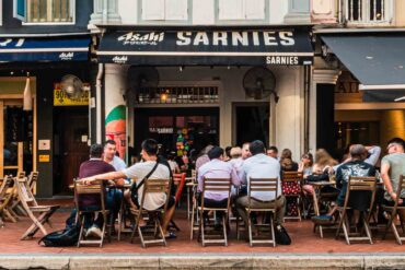 Lively outdoor dining scene at Sarnies café on Telok Ayer Street, Singapore, with diners seated at wooden tables against a backdrop of heritage shophouses.