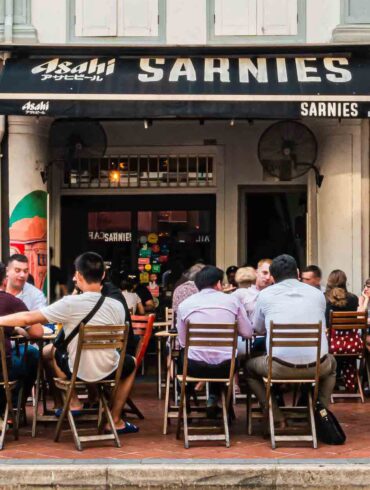Lively outdoor dining scene at Sarnies café on Telok Ayer Street, Singapore, with diners seated at wooden tables against a backdrop of heritage shophouses.