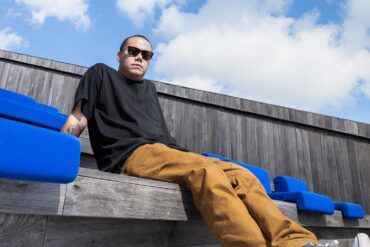 an image of archie dennis lounging at an outdoor bench, with sky and clouds above him, in his background.