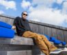 an image of archie dennis lounging at an outdoor bench, with sky and clouds above him, in his background.