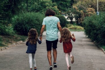 a photo of the father holding the hands of his two daughters, looking joyful from the back.