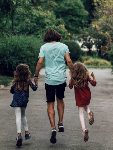 a photo of the father holding the hands of his two daughters, looking joyful from the back.