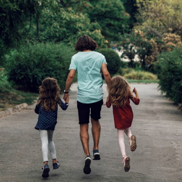a photo of the father holding the hands of his two daughters, looking joyful from the back.