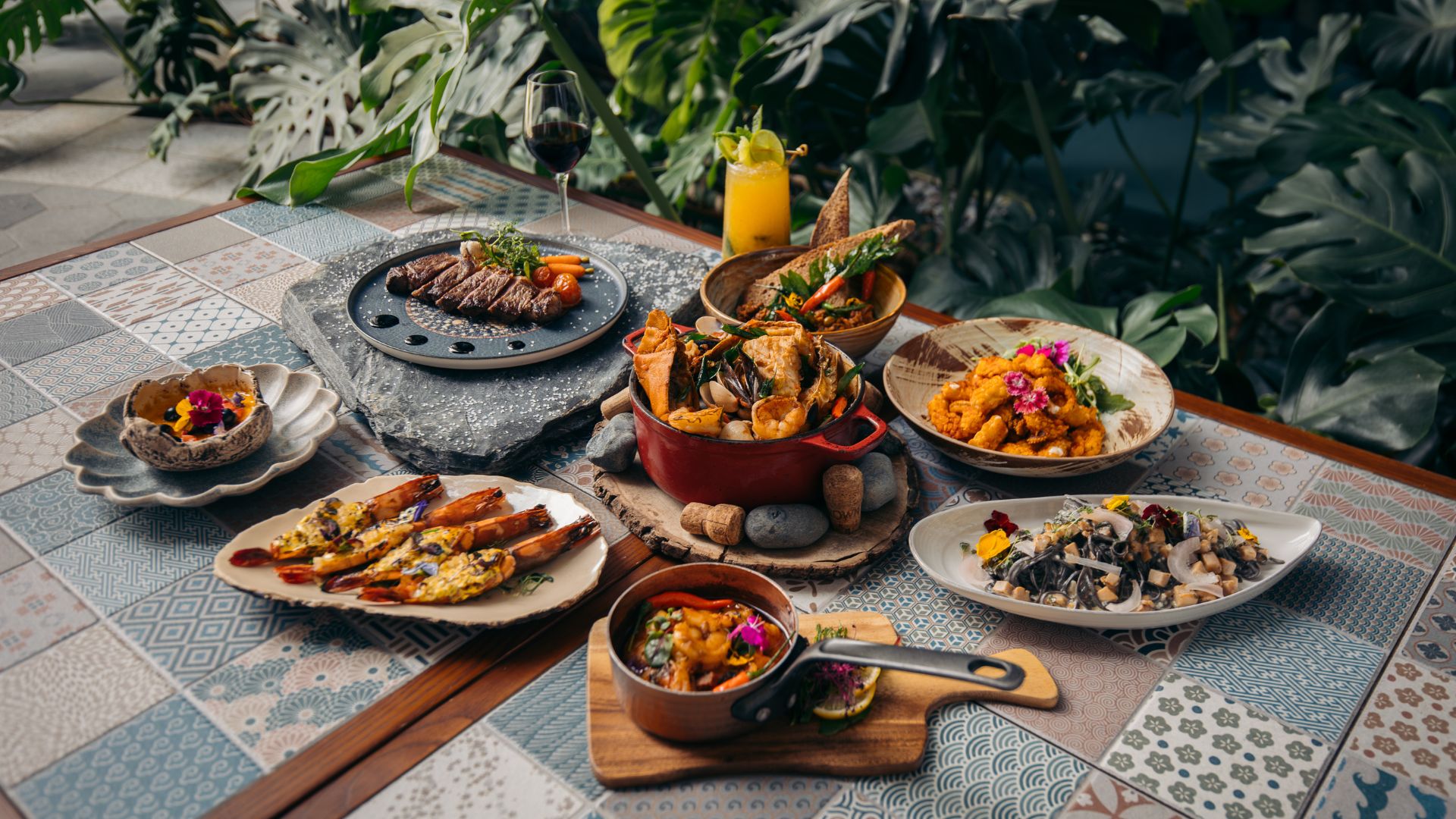 Alfresco dining spread at Winestone Restaurant, Mercure Singapore on Stevens, featuring grilled meats, seafood, sharing plates and wine set against lush tropical greenery.