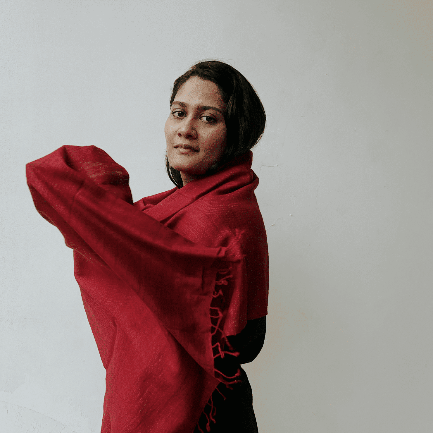 Woman with dark hair wearing a red shawl over a black outfit, standing against a plain white background, looking confidently at the camer