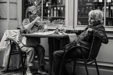 Two elderly women dining al fresco at a European-style cafe with wine glasses and coffee cups on the table – black and white photo capturing candid moments of conversation and leisure.