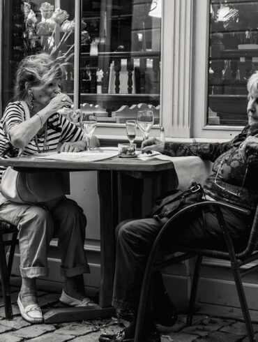 Two elderly women dining al fresco at a European-style cafe with wine glasses and coffee cups on the table – black and white photo capturing candid moments of conversation and leisure.