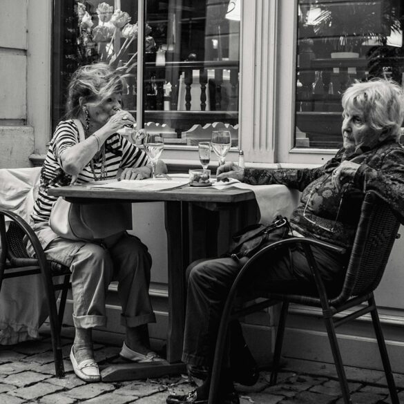 Two elderly women dining al fresco at a European-style cafe with wine glasses and coffee cups on the table – black and white photo capturing candid moments of conversation and leisure.