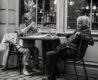 Two elderly women dining al fresco at a European-style cafe with wine glasses and coffee cups on the table – black and white photo capturing candid moments of conversation and leisure.