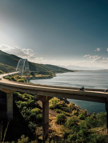 Motorcyclists riding across a scenic curved bridge over a large lake surrounded by green mountains under a bright afternoon sun in Arizona.