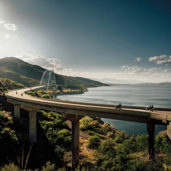 Motorcyclists riding across a scenic curved bridge over a large lake surrounded by green mountains under a bright afternoon sun in Arizona.