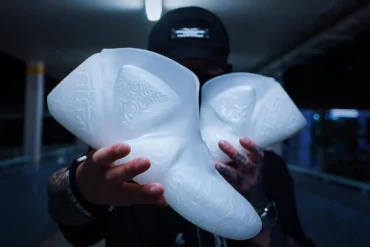 A male model wearing streetwear, holding a pair of 3D-Printed shoes in front of his face.