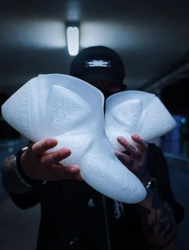 A male model wearing streetwear, holding a pair of 3D-Printed shoes in front of his face.