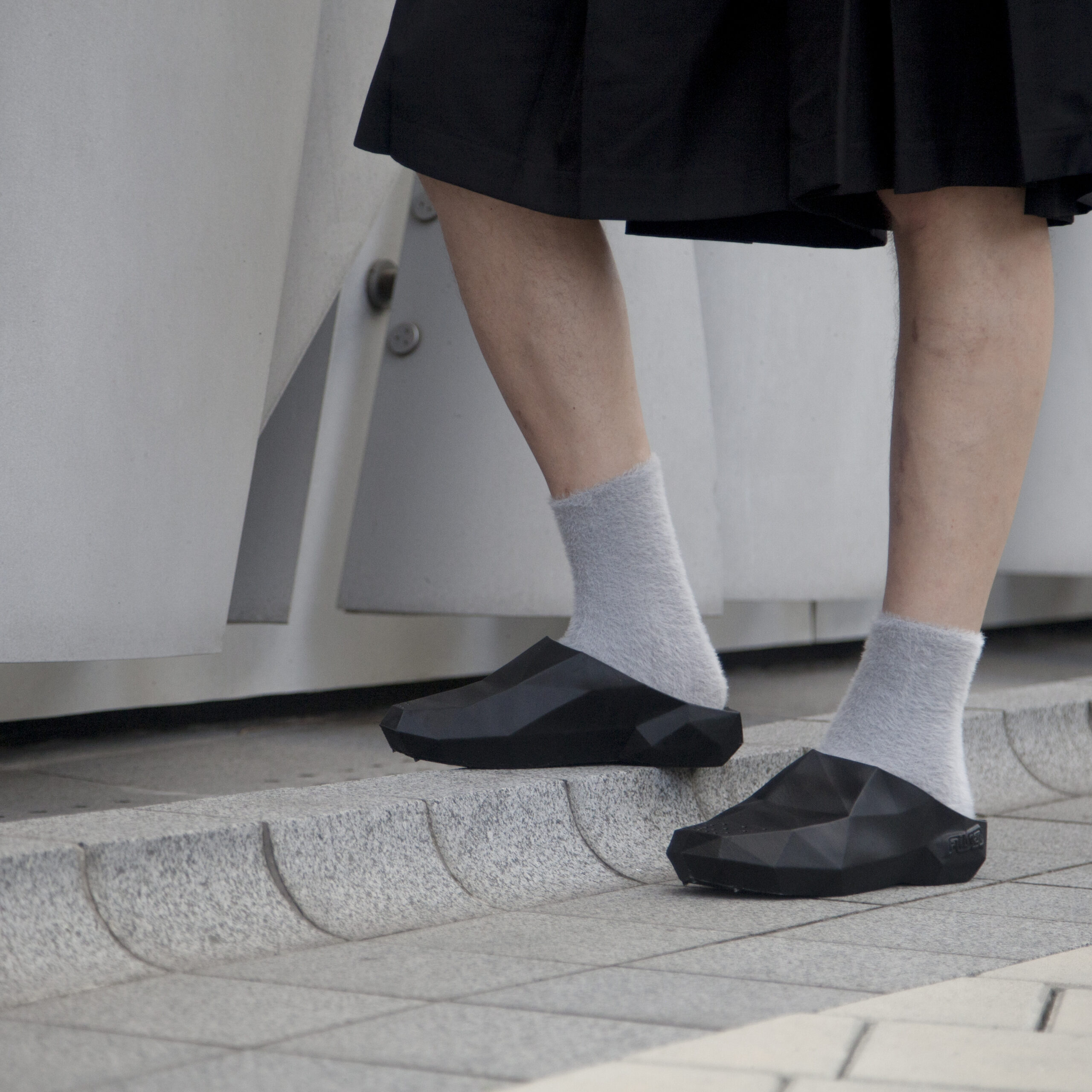 A man wearing a black skirt, black slippers and grey socks on an urban concrete background
