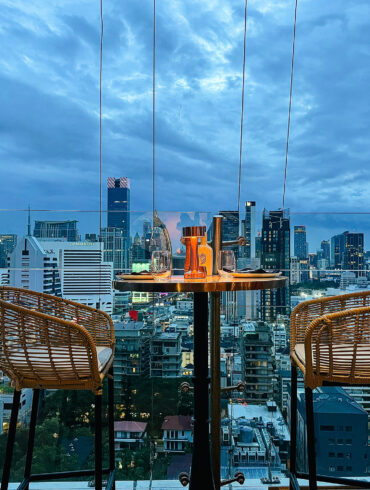 Rooftop table at Pastel Bangkok with rattan chairs overlooking the city skyline at dusk, capturing the Mediterranean-inspired atmosphere from above.