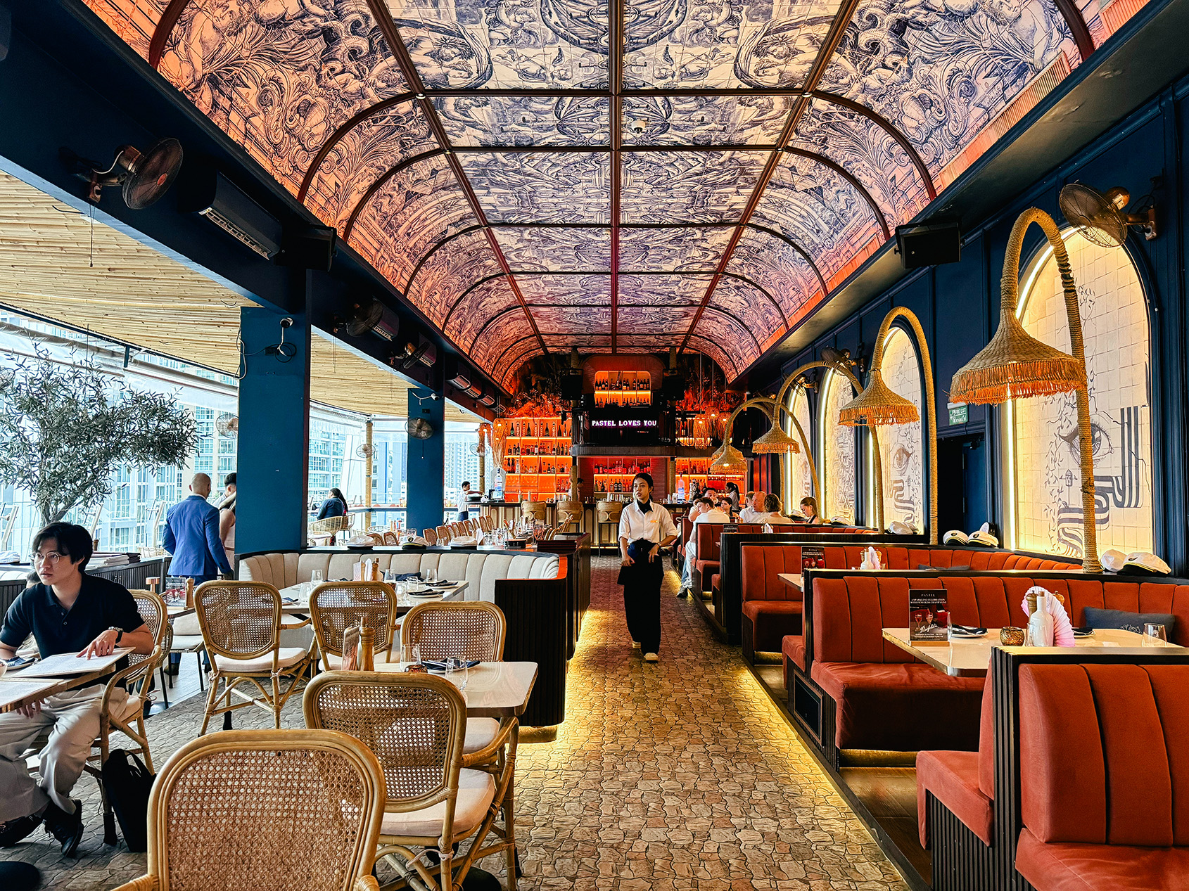 Inside Pastel Bangkok rooftop restaurant with Mediterranean-meets-Bali design, arched ceilings with artistic prints, warm lighting, rattan chairs, and red booth seating.