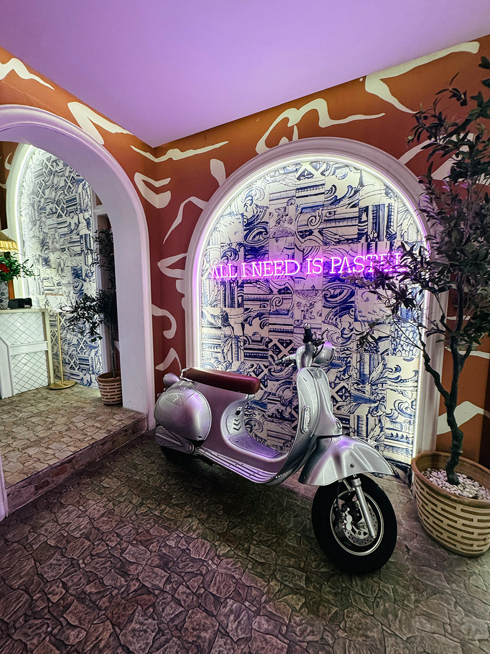 Pastel Bangkok rooftop entrance with a silver Vespa, Mediterranean-inspired blue and white tiles, and neon sign reading “All I Need Is Pastel.”