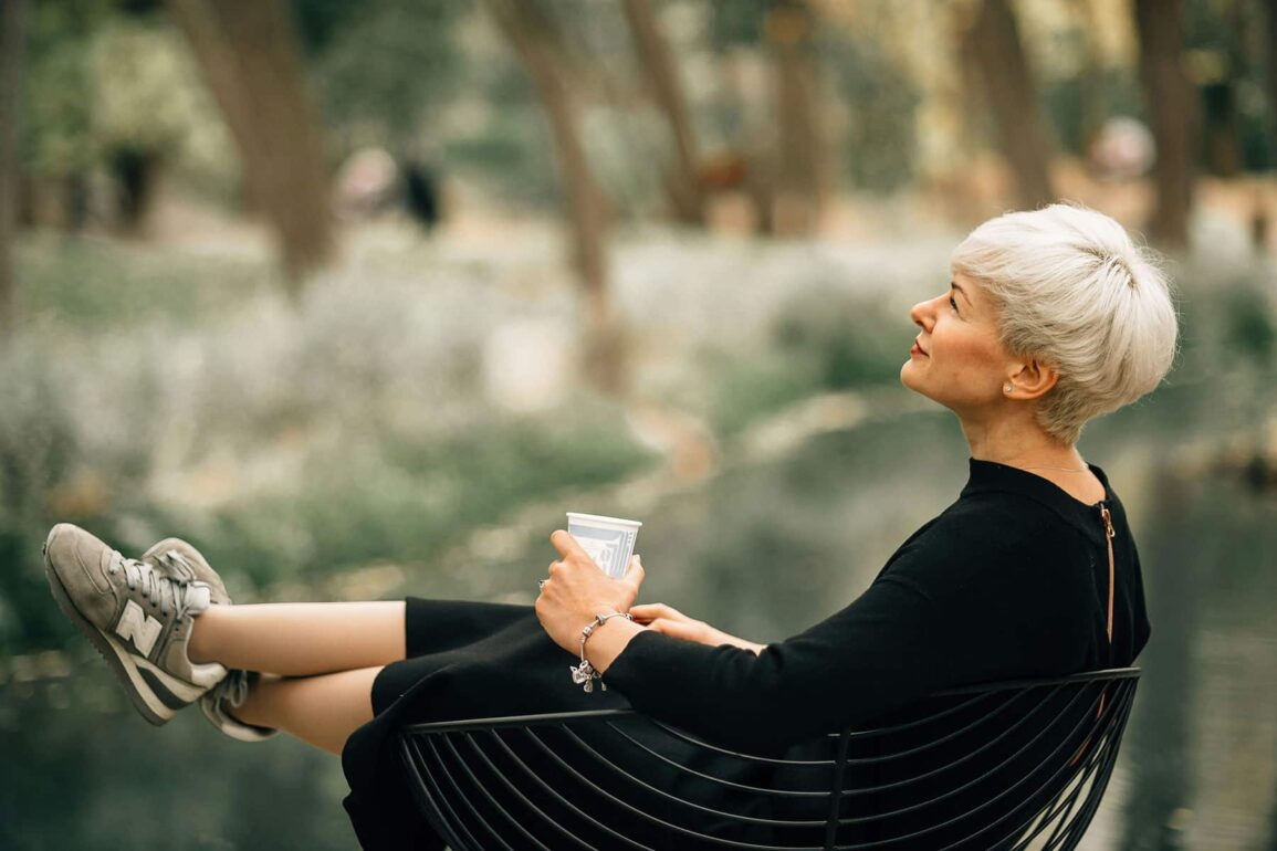 Confident older woman in black dress relaxing outdoors with a coffee, embodying grace, self-assurance, and healthy aging during menopause.