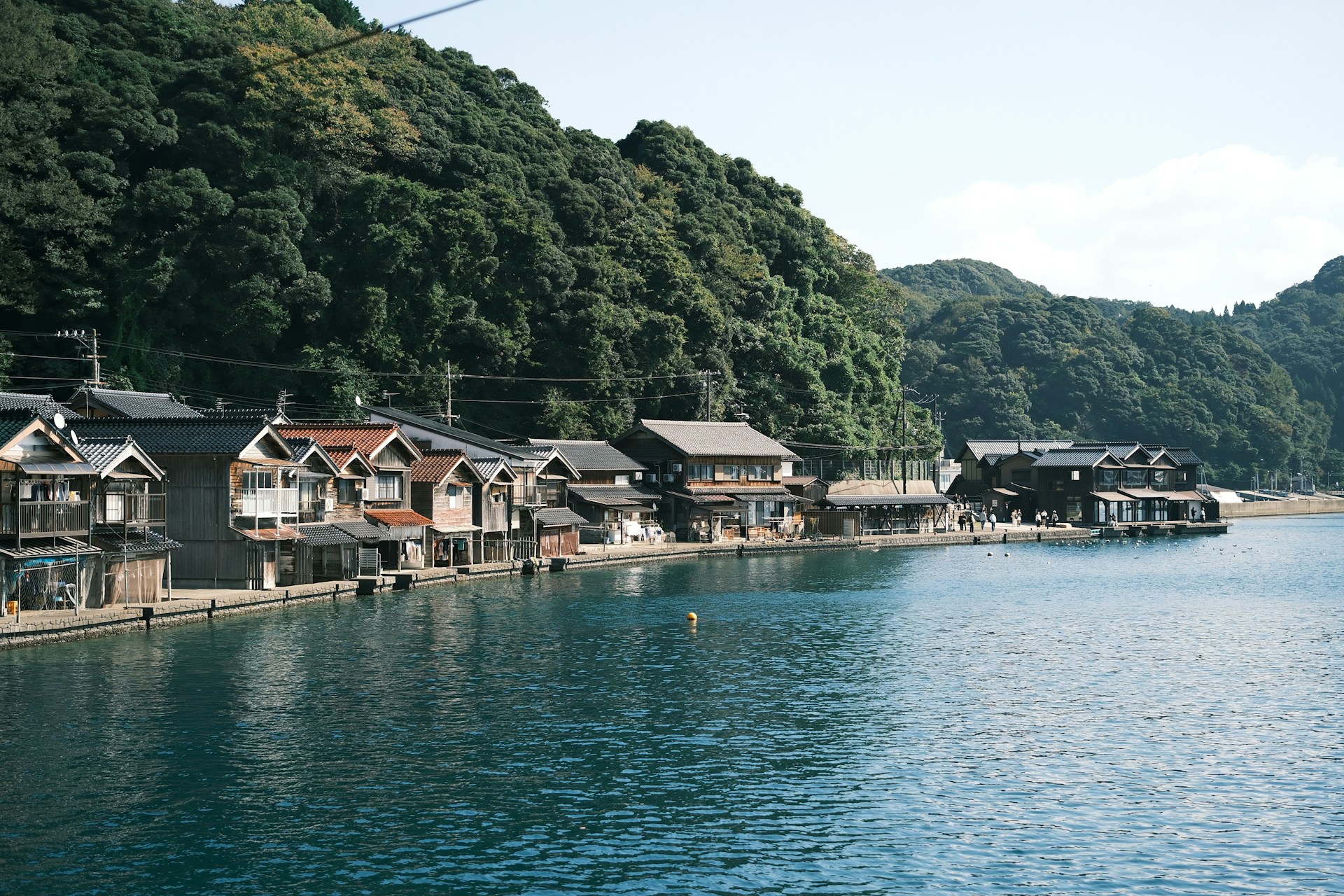 Traditional funaya boathouses lining the waterfront of Ine Bay in Kyoto Prefecture, Japan, with wooden houses built directly over the water against a backdrop of forested hills.