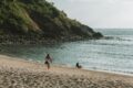 a surfer walking into the waters at West Nusa Tenggara, Indonesia with a surfboard on his side and a girl sitting on the seashore looking out in the horizon.